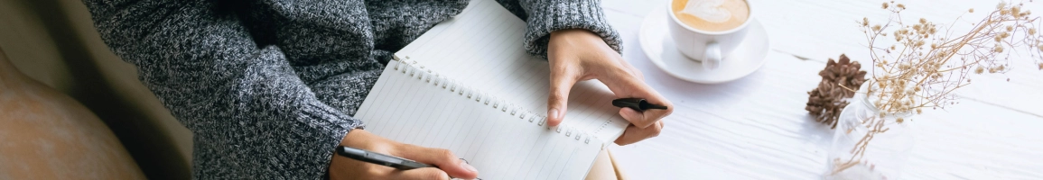 A woman writes in a notebook, with a cappuccino on her desk